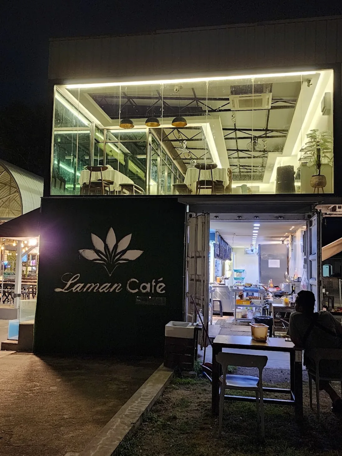 Bahagian hadapan Laman Café pada waktu malam — kontena hitam dengan logo teratai putih, dapur terbuka, dan ruang majlis berdinding kaca di tingkat atas.